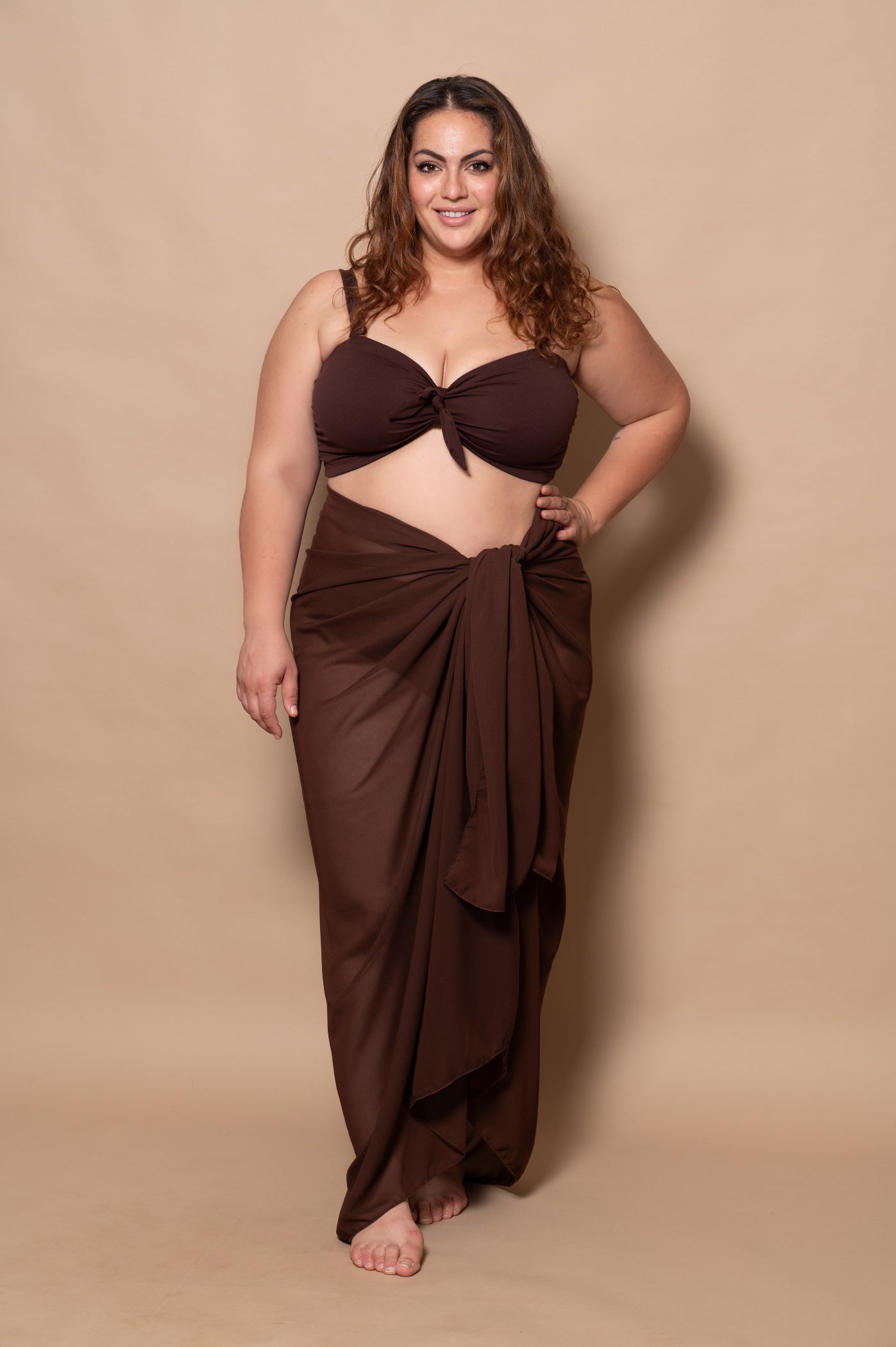 plussize woman wearing a chocolate brown bikini set and pareo cover up against a beige background