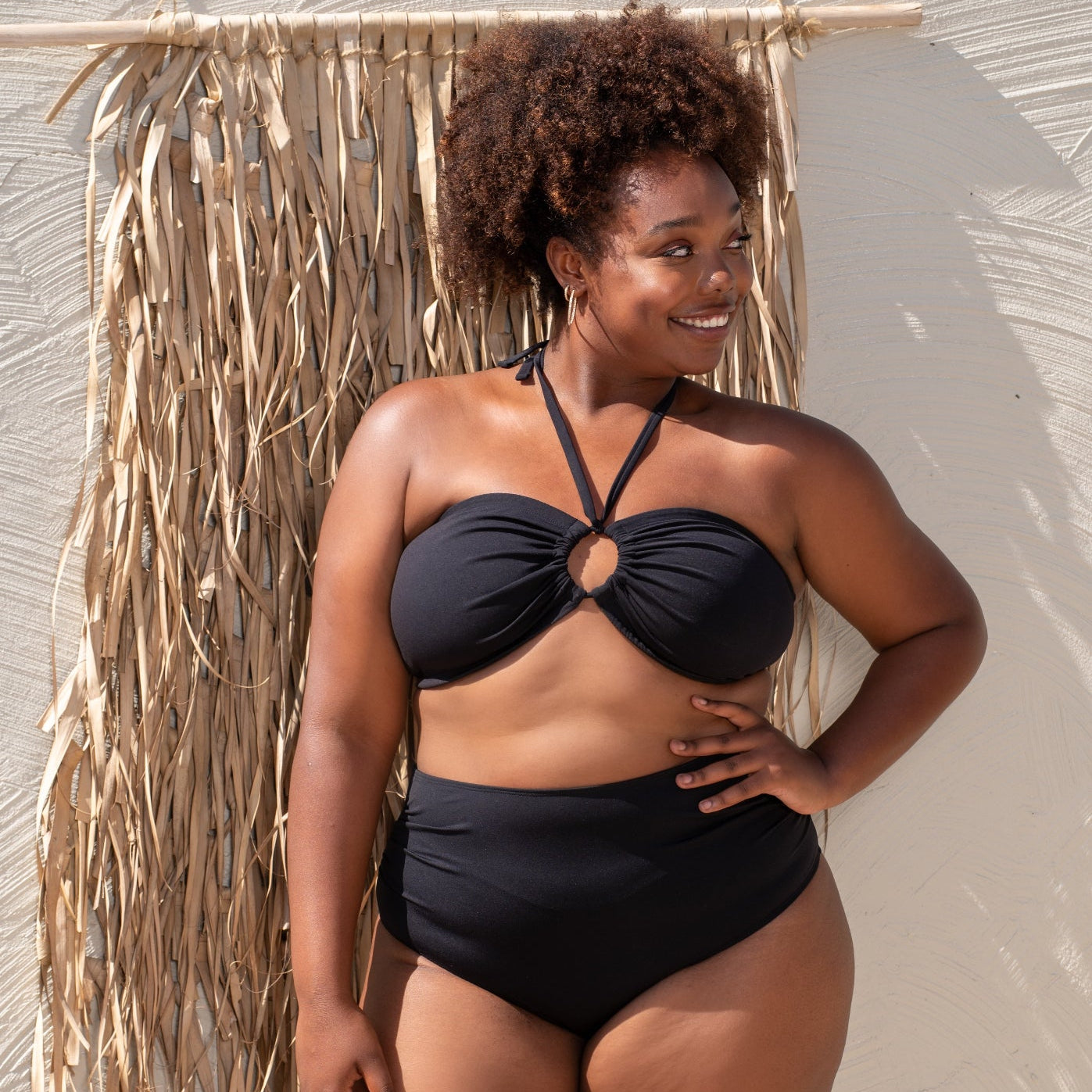 plussize woman wearing a black bikini standing against a textured wall with dried plants.