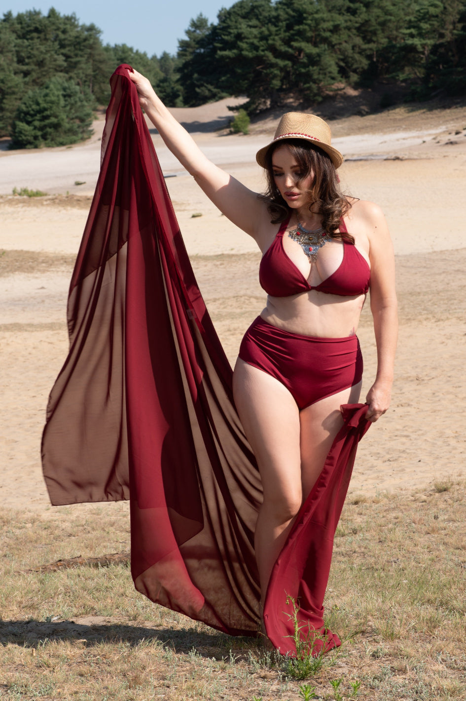 Woman in a wine red bikini holding a wine red pareo coverup in an open area with trees in the background