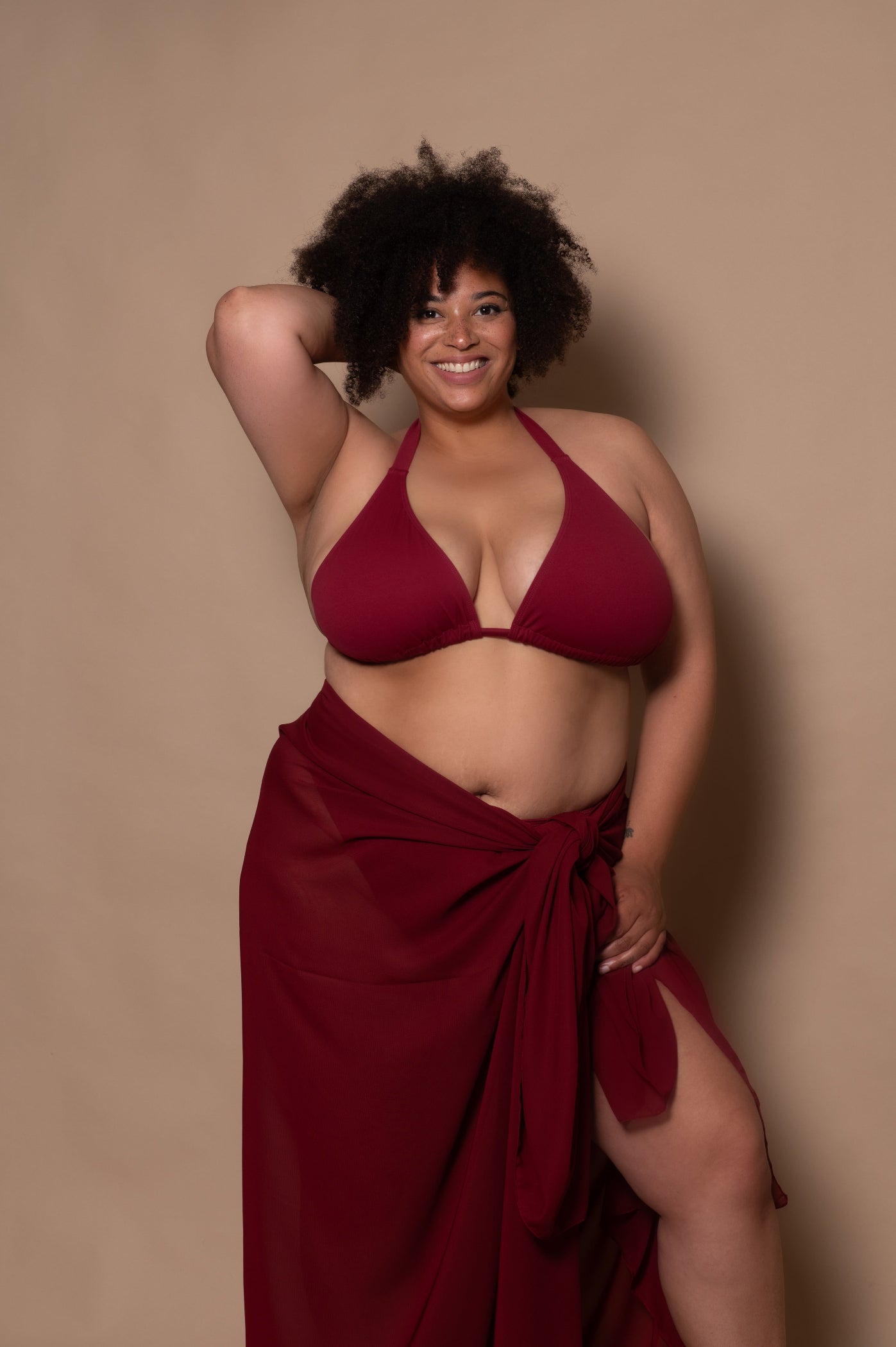 plussize woman wearing a wine red bikini and pareo against a beige background