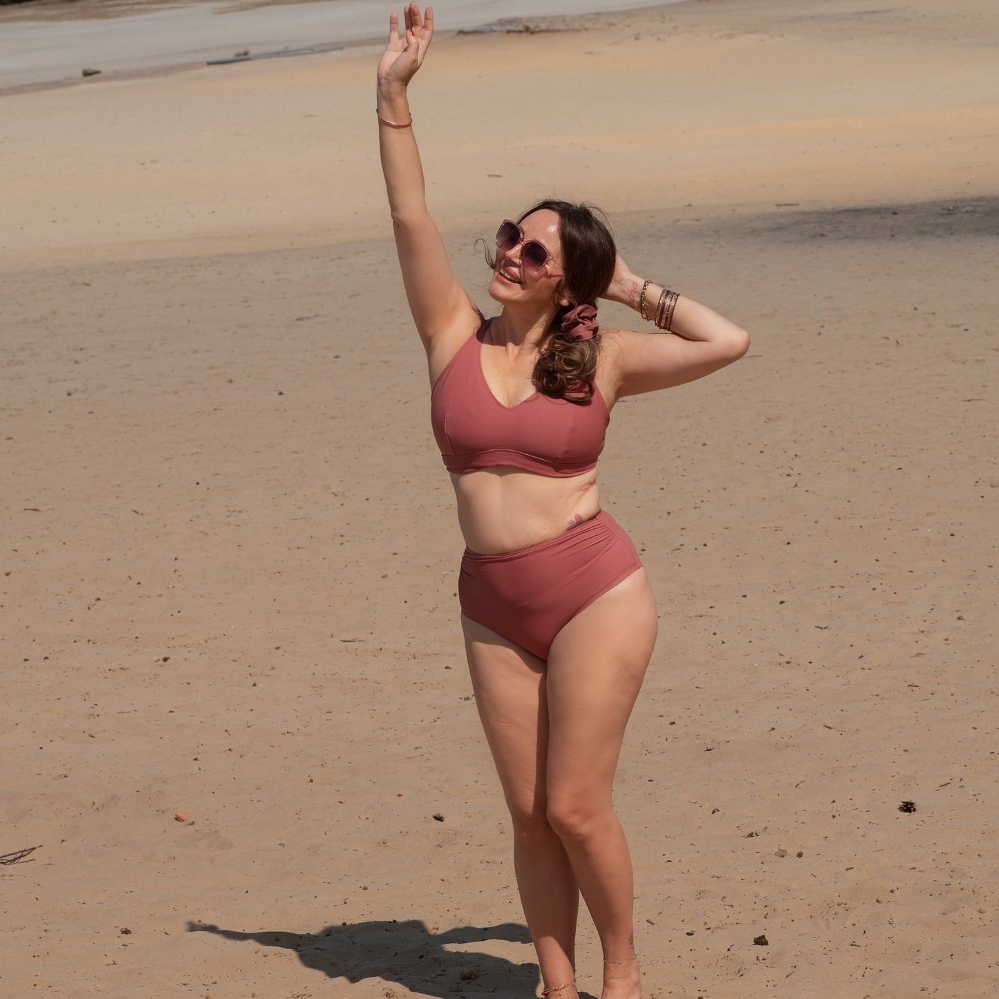 Woman in a ribbed pink bikini and scrunchie standing on a sandy beach with arms raised.