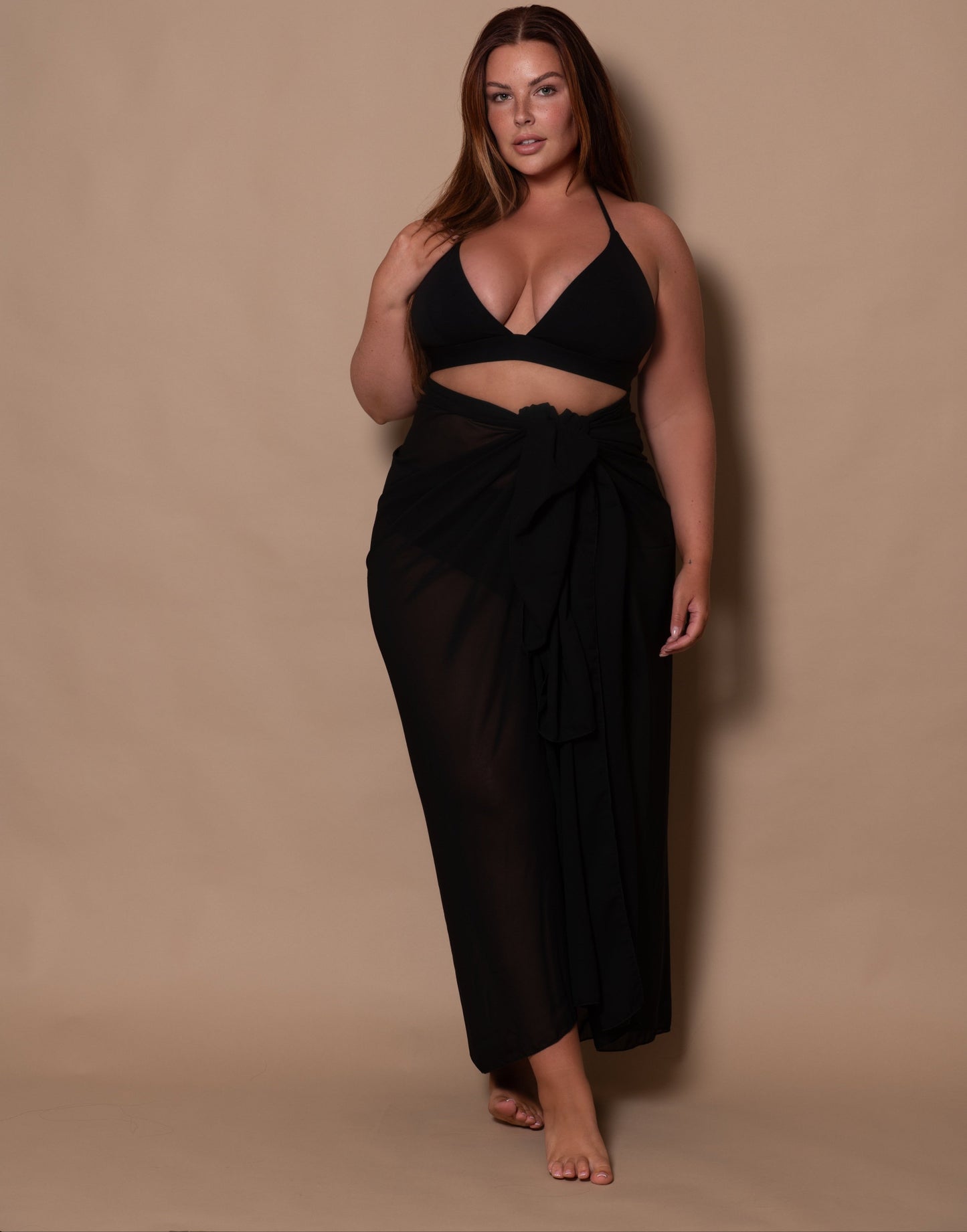plussize woman wearing a black bikini top and a black sarong against a brown background