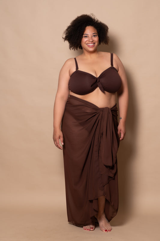 plussize woman wearing a chocolate brown bikini set and pareo cover up against a beige background