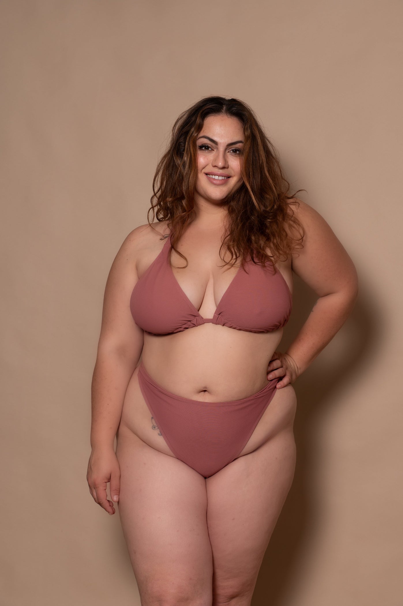 curvy woman wearing a pink rosewood bikini standing against a textured wall.
