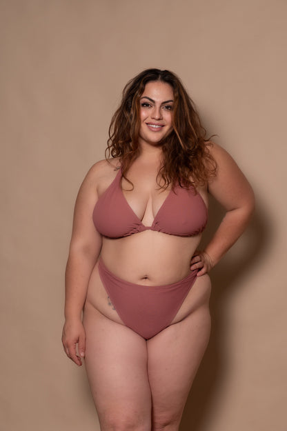 curvy woman wearing a pink rosewood bikini standing against a textured wall.