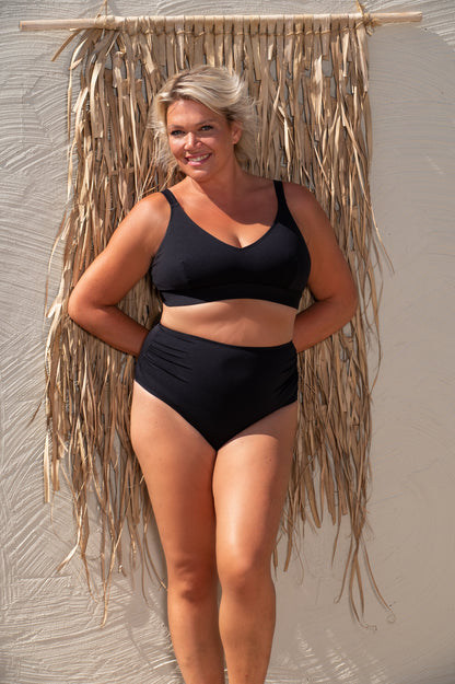 Woman wearing a black bikini standing against a textured wall with dried plants.