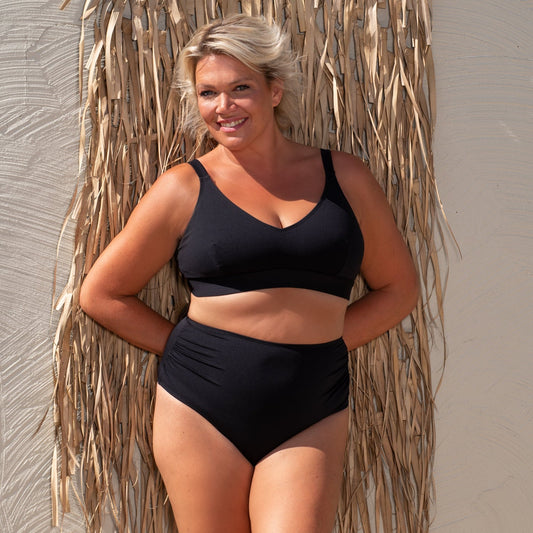 Woman wearing a black bikini standing against a textured wall with dried plants.