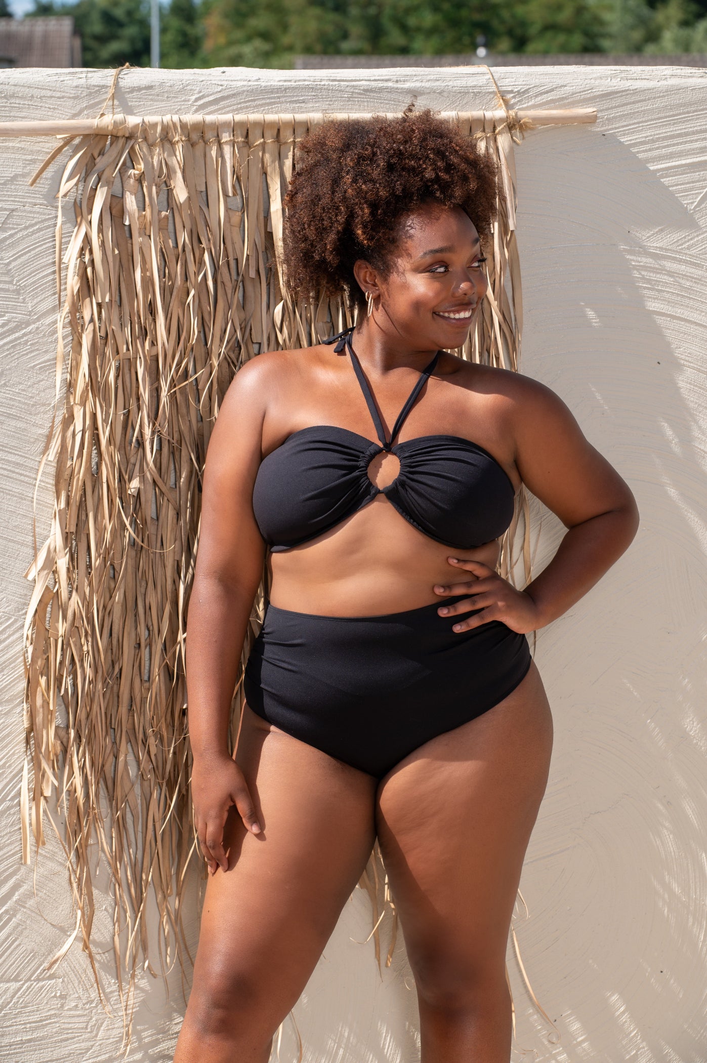 plussize woman wearing a black bikini standing against a textured wall with dried plants.