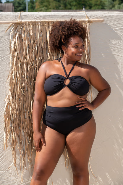 plussize woman wearing a black bikini standing against a textured wall with dried plants.
