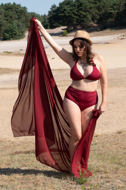 Woman in a wine red bikini holding a wine red pareo coverup in an open area with trees in the background