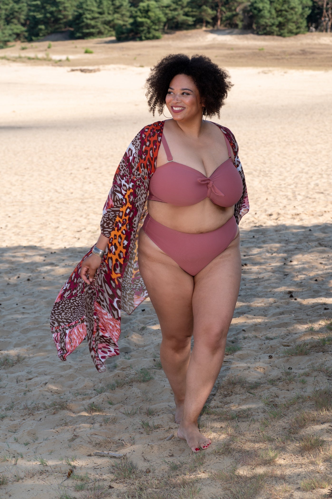 plussize woman in a pink bikini and colorful cover-up walking on a sandy beach.