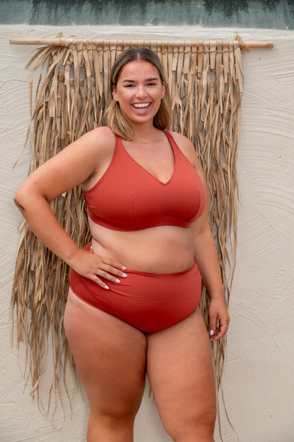 plussize woman wearing a rust-red bikini standing against a textured wall with straw decorations.