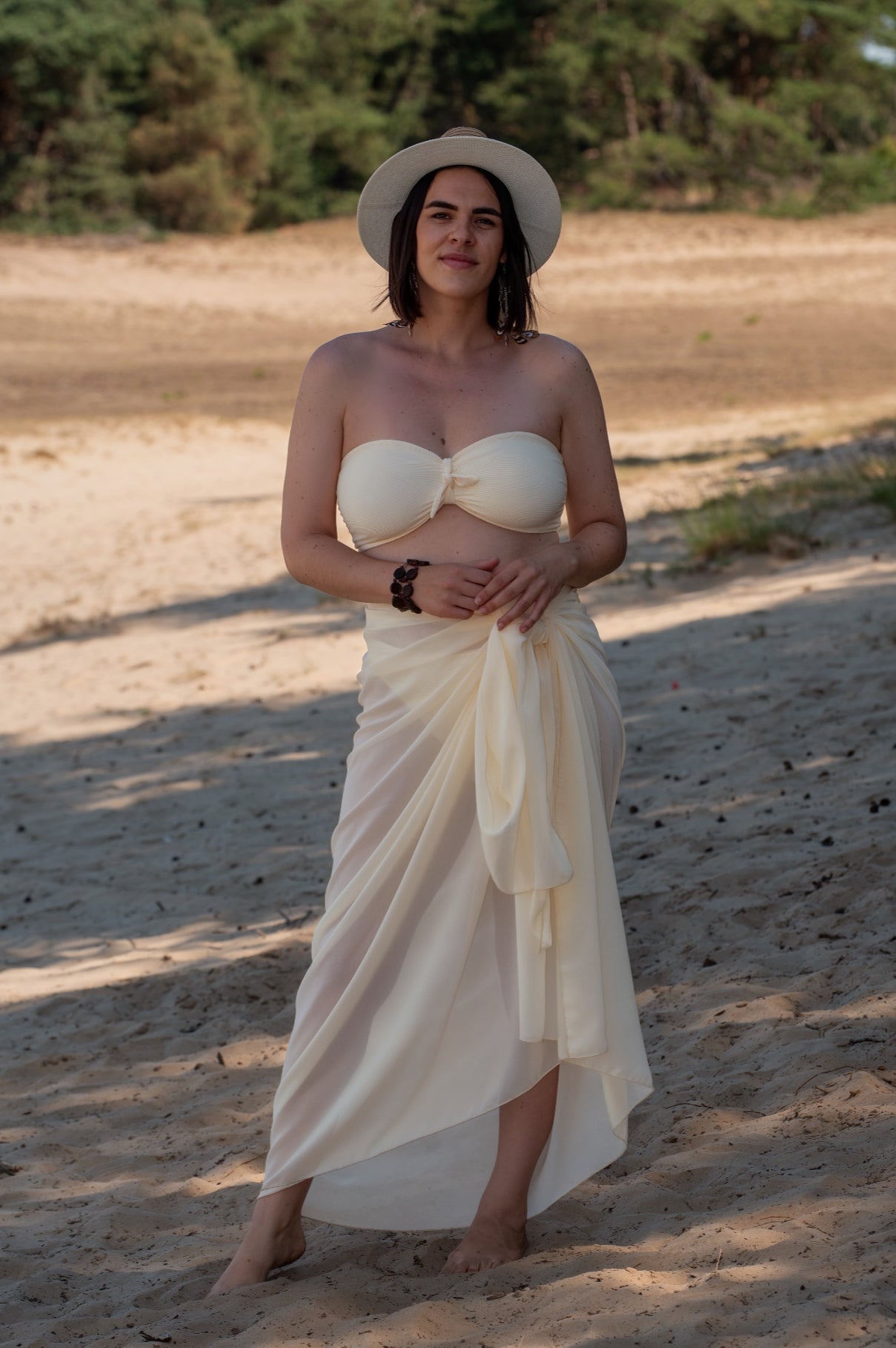 Woman in a white bikini top and pareo standing on a sandy beach with trees in the background
