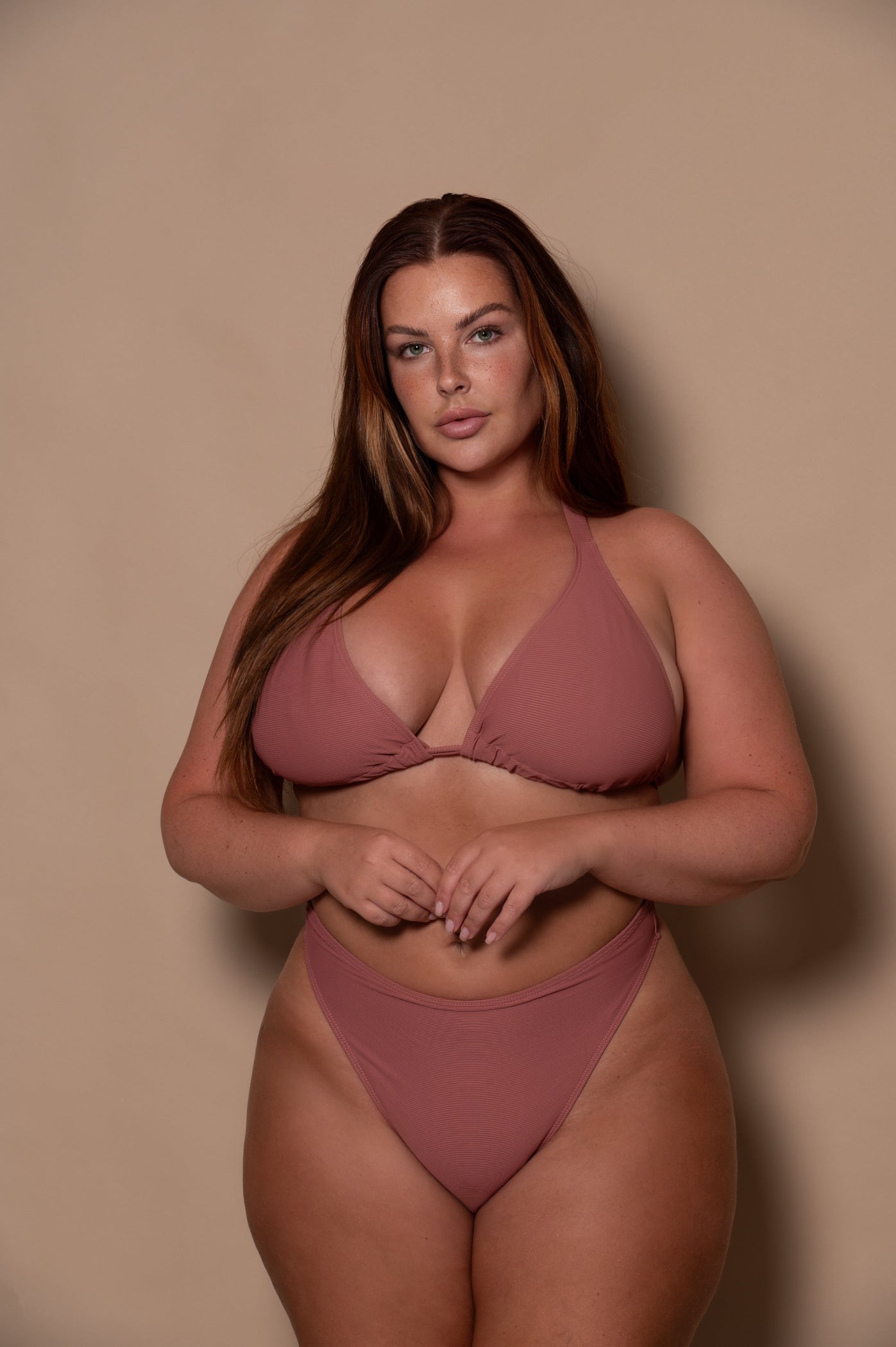 curvy woman wearing a pink rosewood bikini standing against a textured wall.