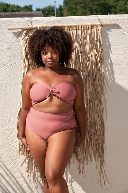 plussize woman in a pink bikini standing against a textured wall with straw decorations.