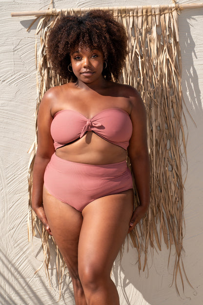 curvy woman wearing a pink rosewood bikini standing against a textured wall.