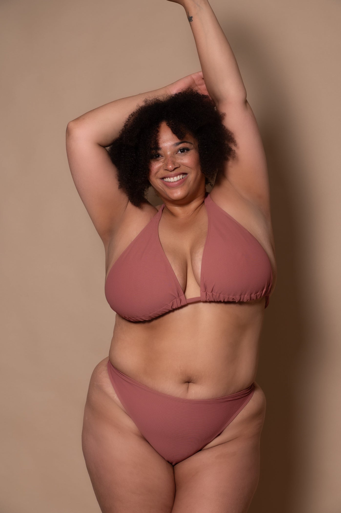 curvy woman wearing a pink rosewood bikini standing against a textured wall.