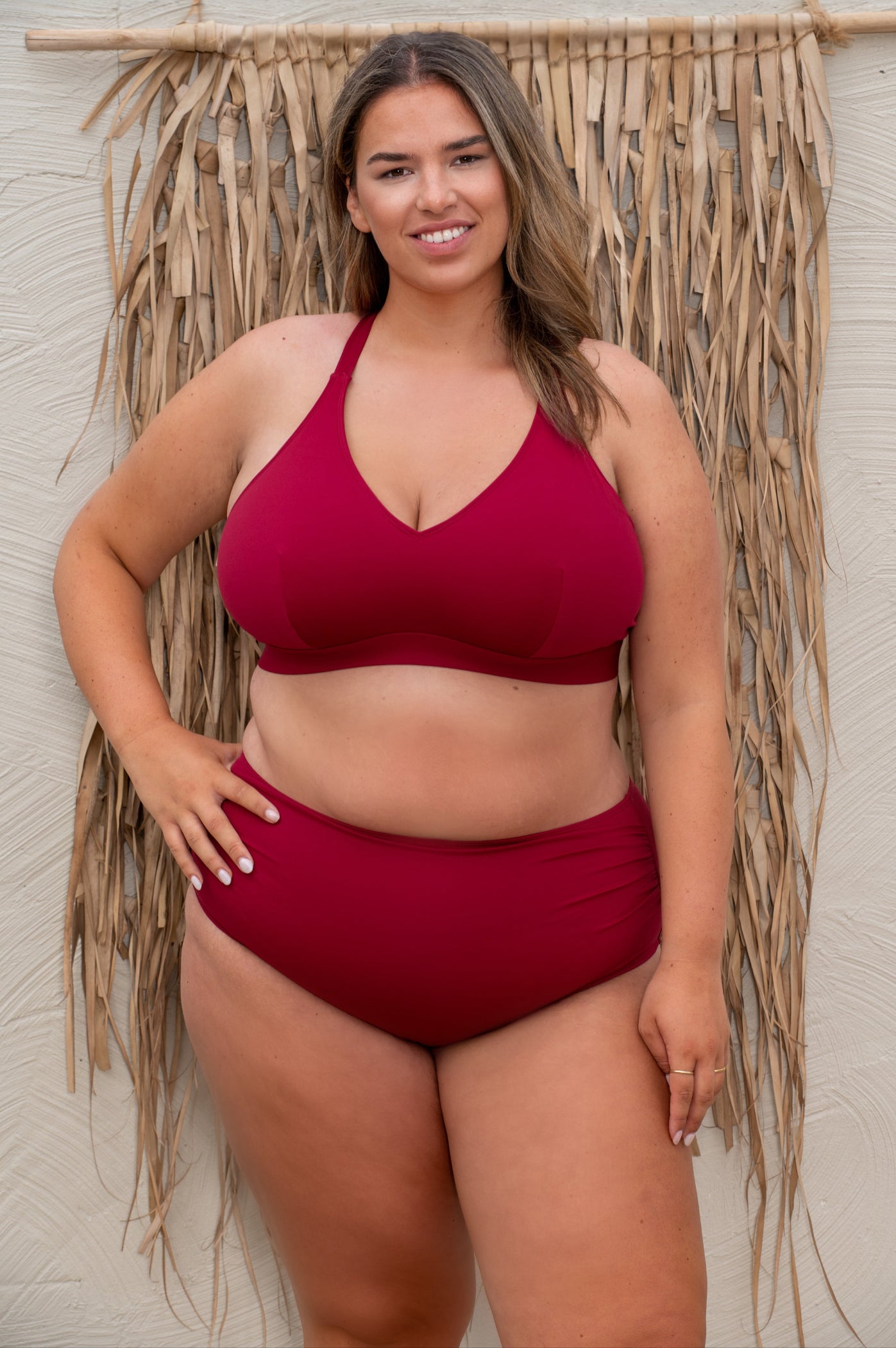 plussize woman wearing a red bikini standing against a textured wall with dried plants.