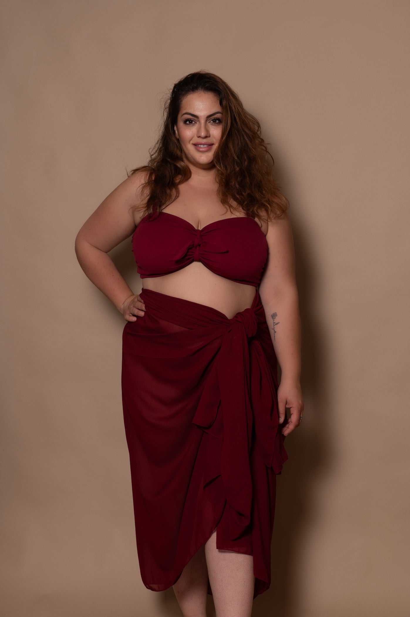 plussize woman wearing a wine red bikini  and pareo cover up against a beige background